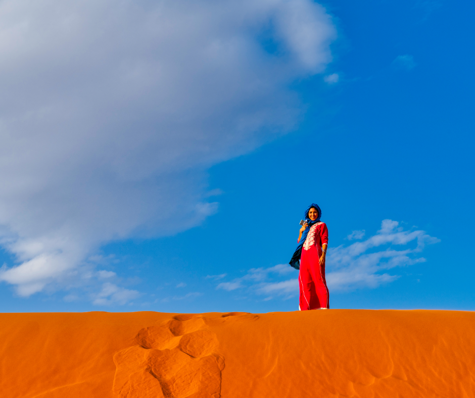 Woman in a red dress and blue headscarf standing on an orange Sahara dune