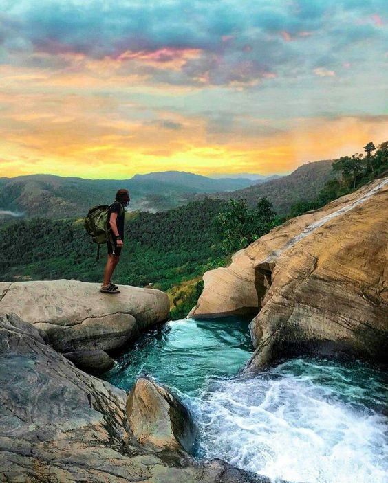 Hiker standing on rocks above a waterfall pool overlooking green hills at sunset