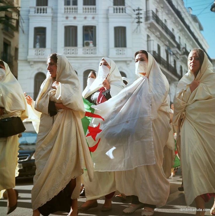 Women in white haik veils marching with an Algerian flag through an Algiers street
