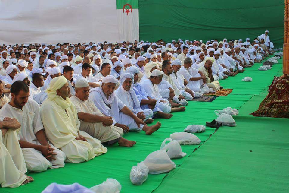 Men in white robes and turbans seated on green mats for Eid prayers