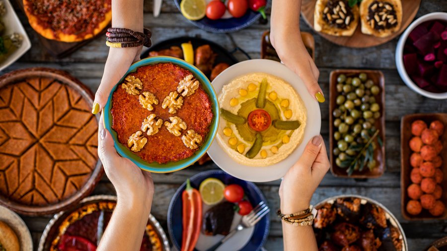 Hands holding plates of hummus and muhammara over a table of mezze dishes