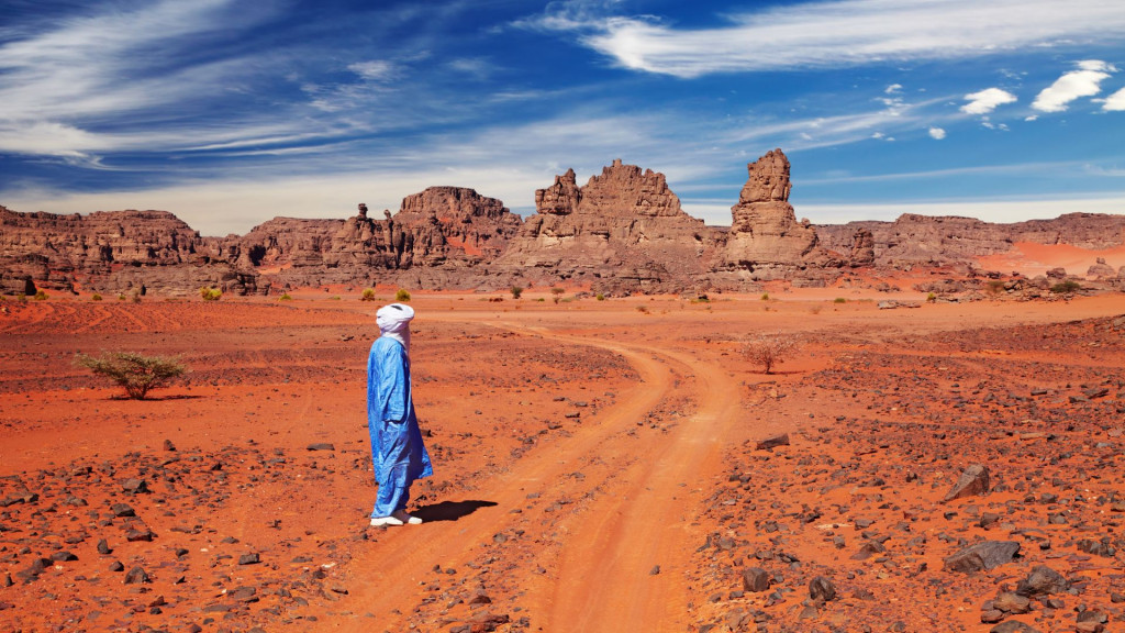 Tuareg man in a blue robe standing on a red desert track near Djanet