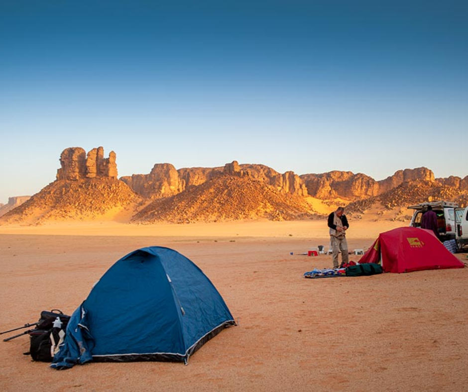 Blue and red tents pitched in orange sand below sandstone rock towers in the Algerian Sahara