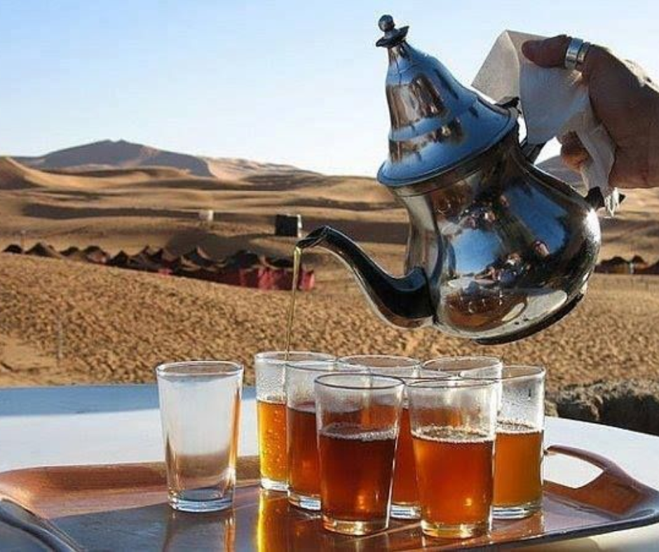 Mint tea poured from a silver teapot into glasses, with desert camp and dunes behind