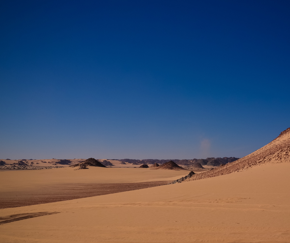 Wide sandy plain with small dark hills and a deep blue sky in the Algerian Sahara
