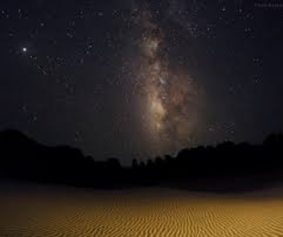 Milky Way arching over a dark horizon and rippled sand dunes at night