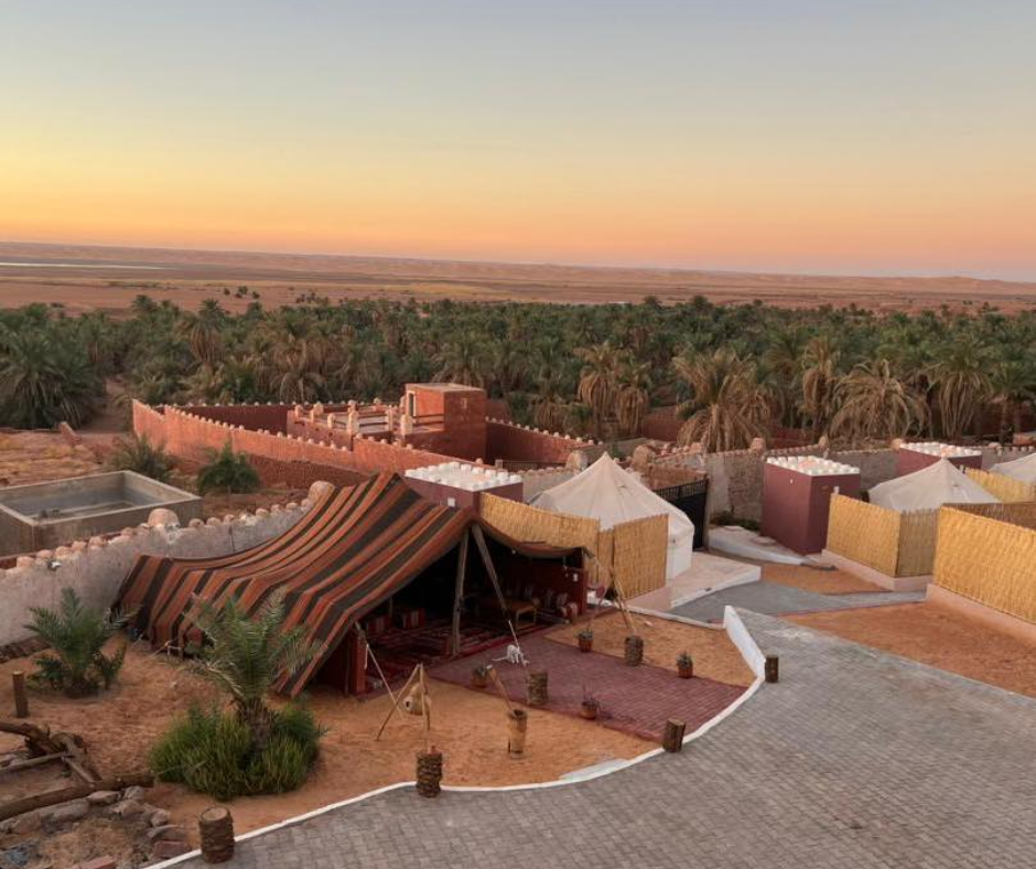 Desert camp with striped tents and walled huts above a palm grove at sunset in Timimoun