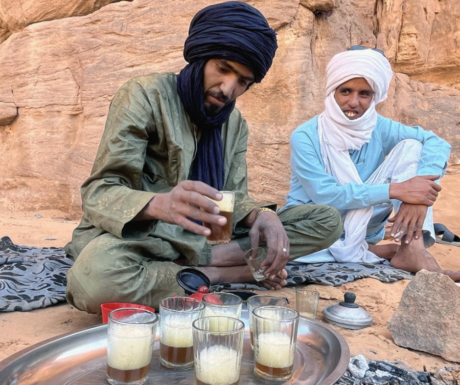 Two Tuareg men in turbans pouring tea into small glasses on a tray beside rock