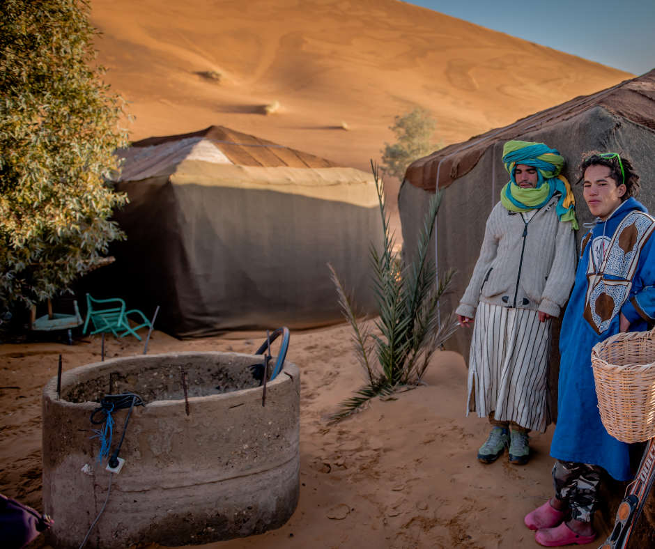 Two men in traditional dress beside Berber tents and a well in the dunes