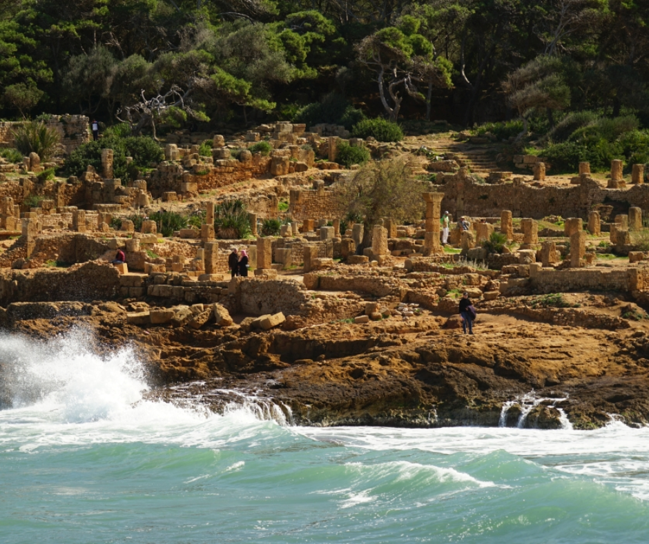 Roman ruins of Tipaza on the rocky shore with waves breaking below pine trees
