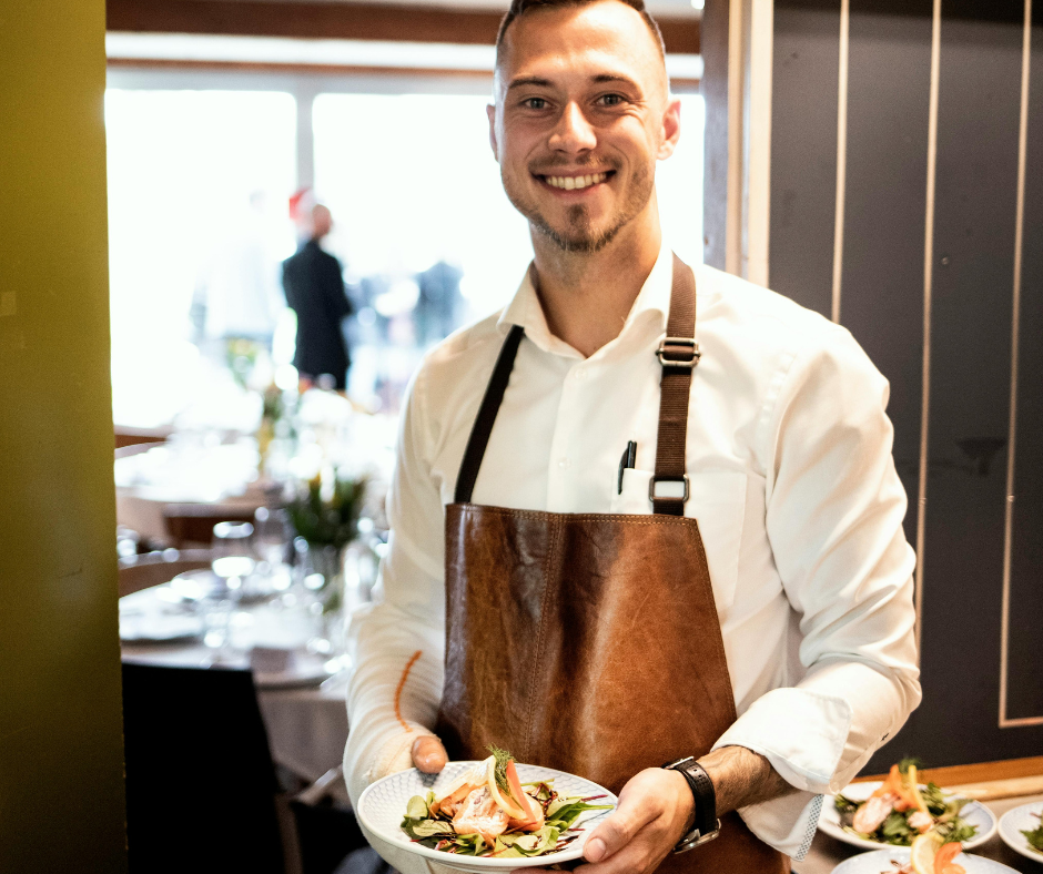 Smiling waiter in a leather apron carrying two plated dishes in a restaurant