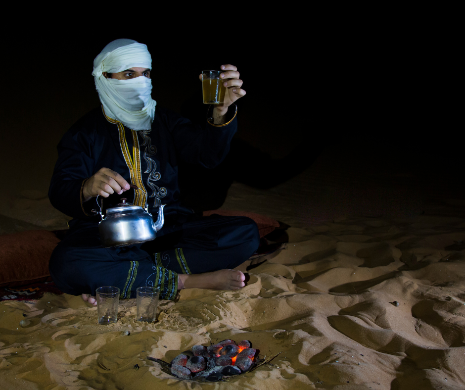 Man in a white veil pouring Saharan tea by a charcoal fire at night