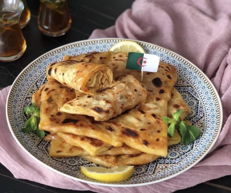 Folded mhajeb crepes on a patterned plate with an Algerian flag and mint tea