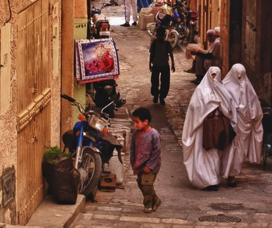 Narrow M'Zab lane with women in white haik, a child and parked motorbikes