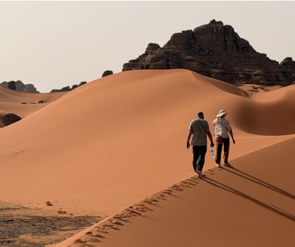 Two hikers walking along a dune ridge toward rock formations in the Sahara