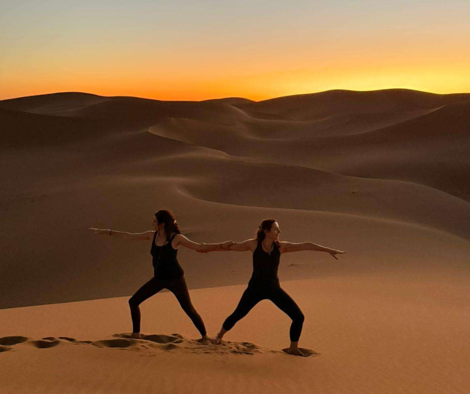 Two women doing yoga warrior pose on a dune at sunset