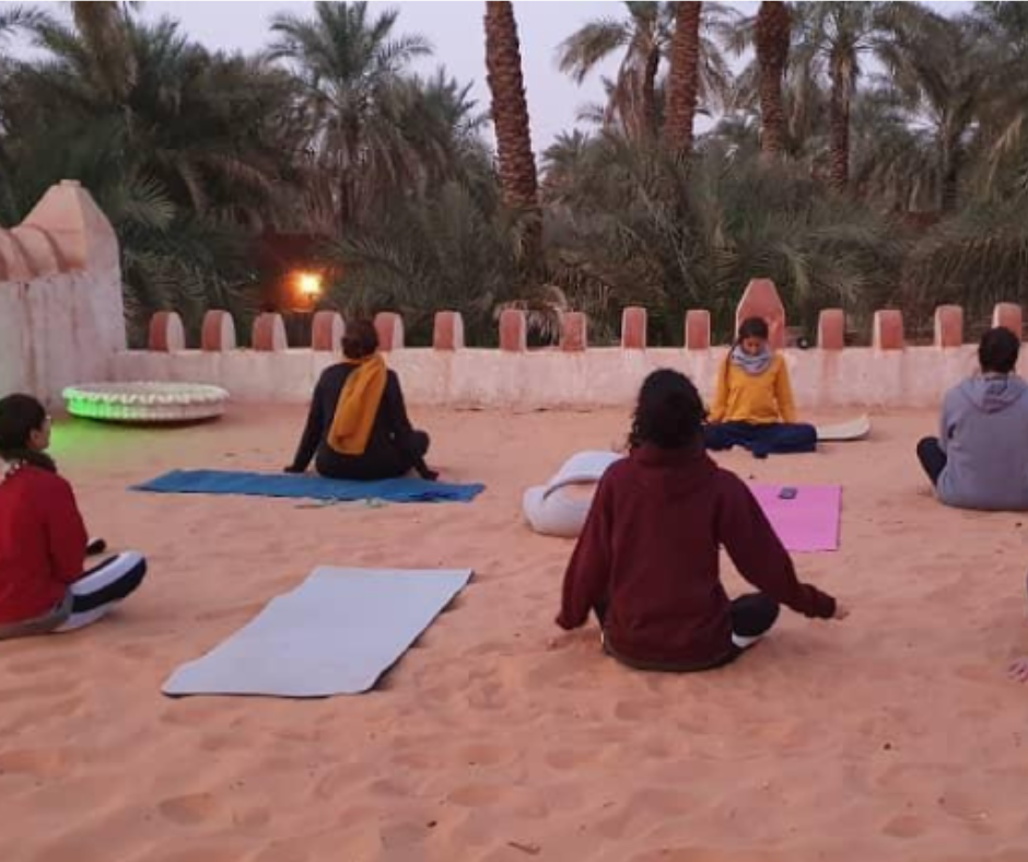 Group sitting on yoga mats on sand in a palm grove courtyard