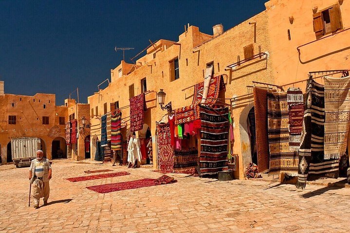 Elderly man with a cane crossing a market square hung with woven carpets in the M'Zab