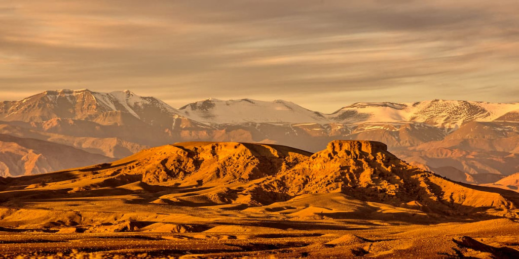 Snow-capped mountains rising behind bare ochre hills in evening light